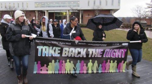 A group of people marches outdoors, holding a banner that reads "TAKE BACK THE NIGHT" during an event for National Crime Victims' Rights Week.